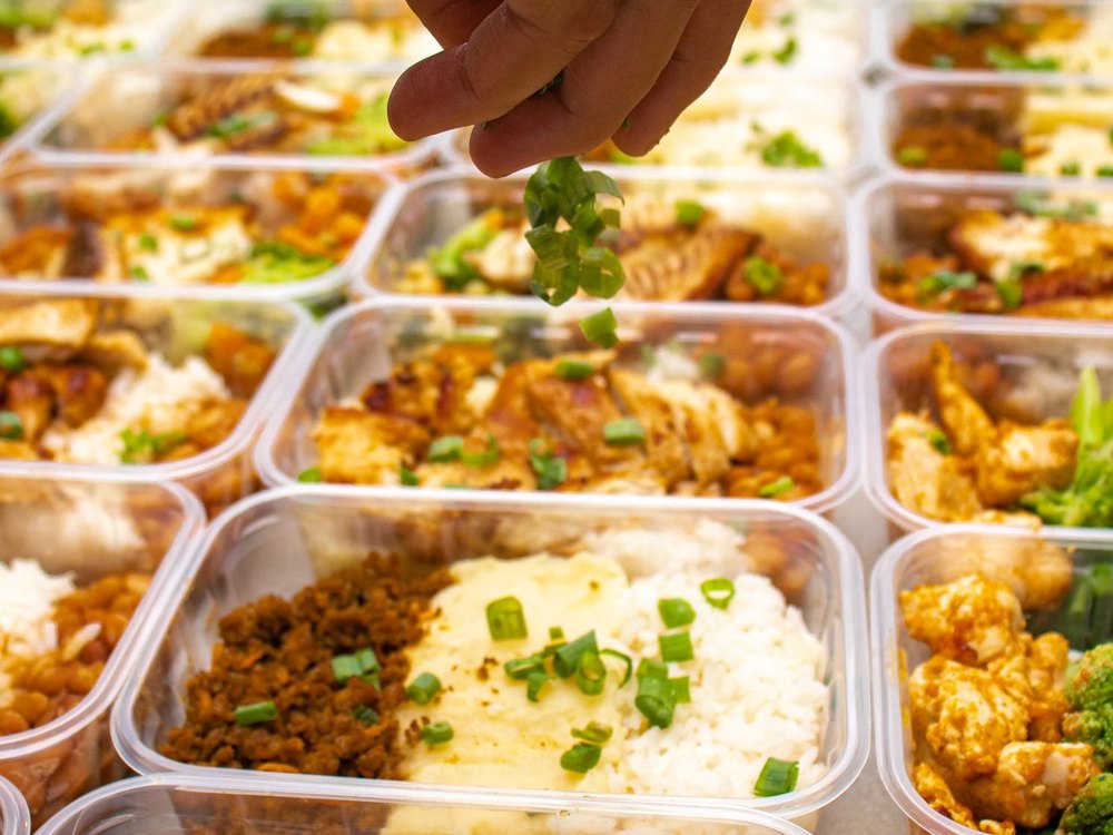 Soul-food meal-prep containers of protein, rice, and vegetables being garnished fresh